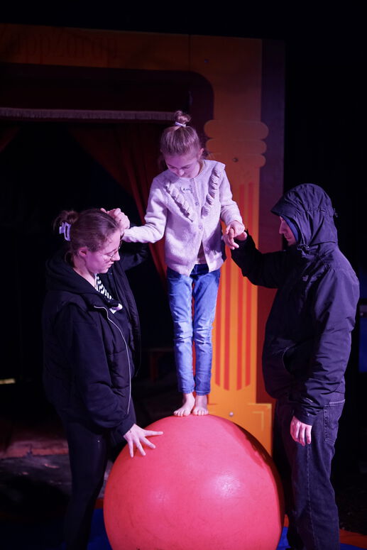 "Ava balanciert im Zirkus auf einem Ball, gesichert von zwei anderen Personen"