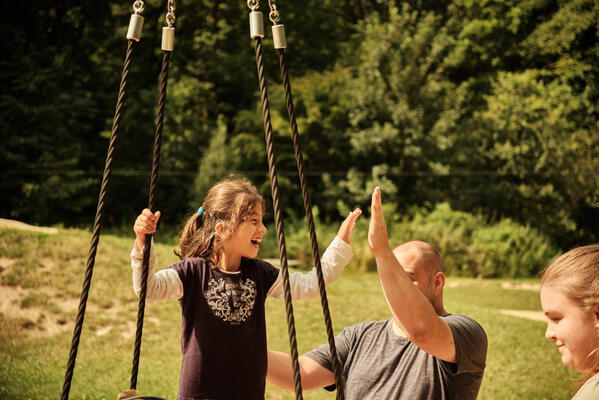Ein lachendes Maedchen auf einer Schaukel gibt einem Erwachsenen freudig ein High-Five, waehrend eine weitere Person daneben steht - eine Szene voller gemeinsamer Freude und Teilhabe in der Natur.